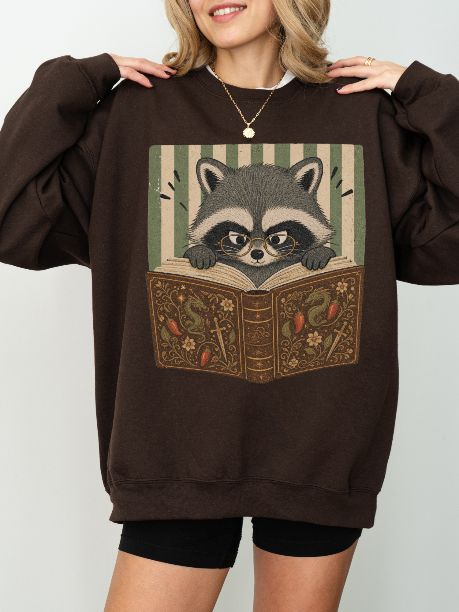 woman wearing dark chocolate Gildan raccoon reading book sweatshirt with bookish raccoon wearing glasses and fantasy book design