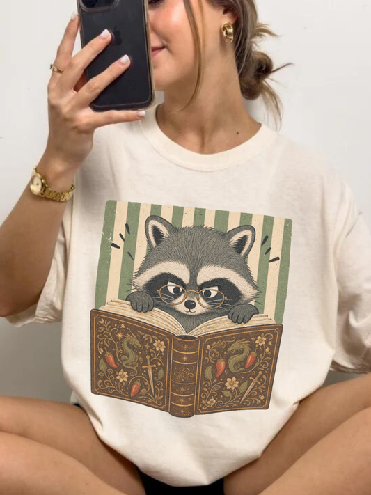 ivory Comfort Colors raccoon reading book t-shirt flat lay featuring a cute raccoon with glasses reading a vintage fantasy book