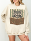 woman wearing sand Gildan raccoon reading book sweatshirt featuring a cute raccoon with glasses and vintage fantasy book