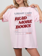 woman wearing a blossom pink Comfort Colors shirt featuring a library card inspired read more books graphic in red lettering, oversized book lover tee styled with biker shorts and gold jewelry