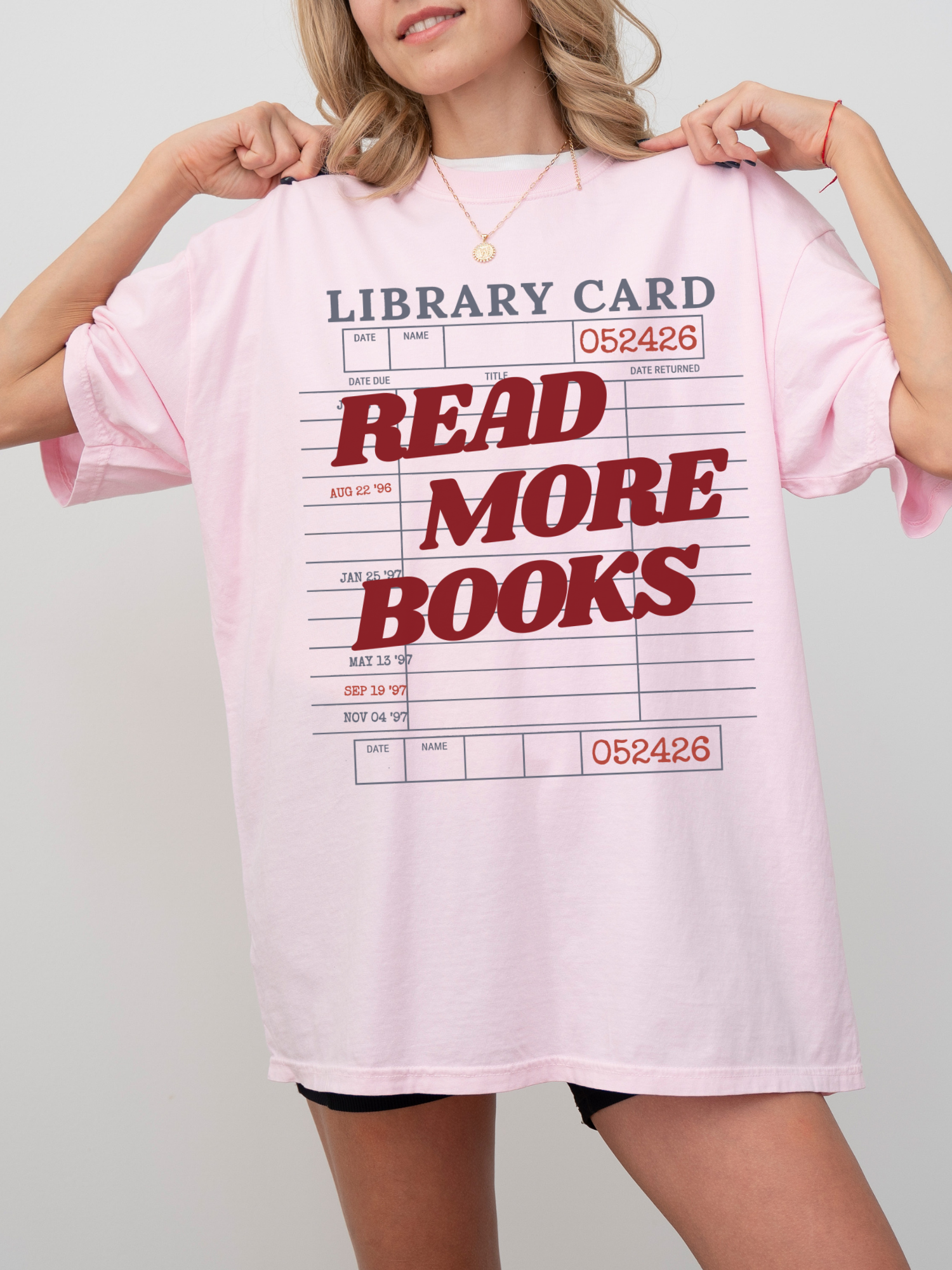 woman wearing a blossom pink Comfort Colors shirt featuring a library card inspired read more books graphic in red lettering, oversized book lover tee styled with biker shorts and gold jewelry