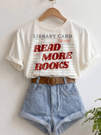 ivory Comfort Colors shirt hanging with rolled sleeves featuring a vintage library card graphic and read more books text in red lettering, styled with denim shorts and a brown belt for a cozy bookish aesthetic