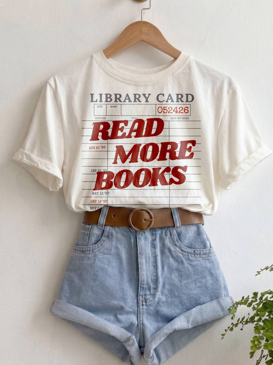 ivory Comfort Colors shirt hanging with rolled sleeves featuring a vintage library card graphic and read more books text in red lettering, styled with denim shorts and a brown belt for a cozy bookish aesthetic