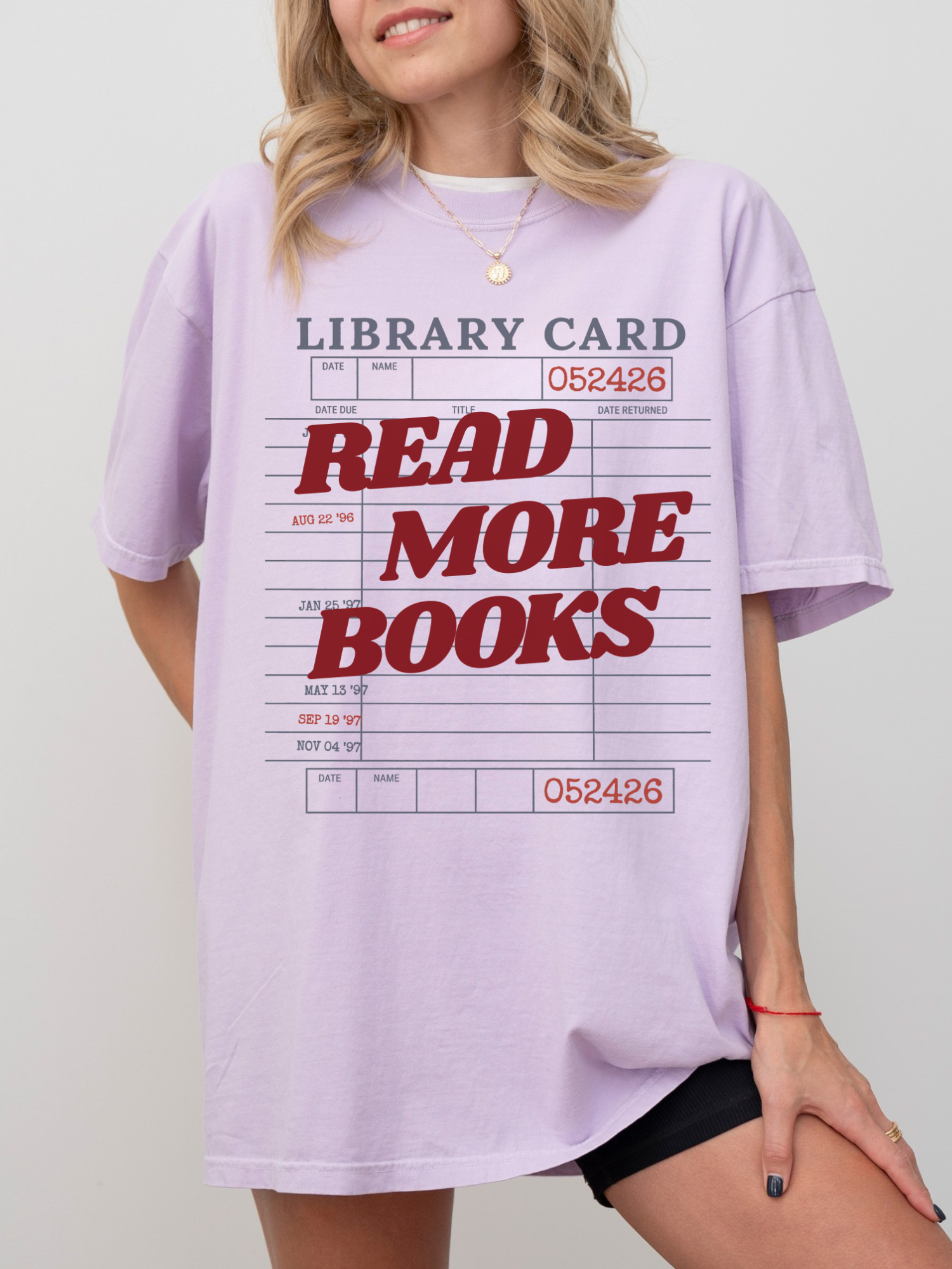 woman wearing an orchid Comfort Colors shirt featuring a library card graphic with read more books text in red lettering, oversized reading shirt styled with biker shorts and delicate gold jewelry