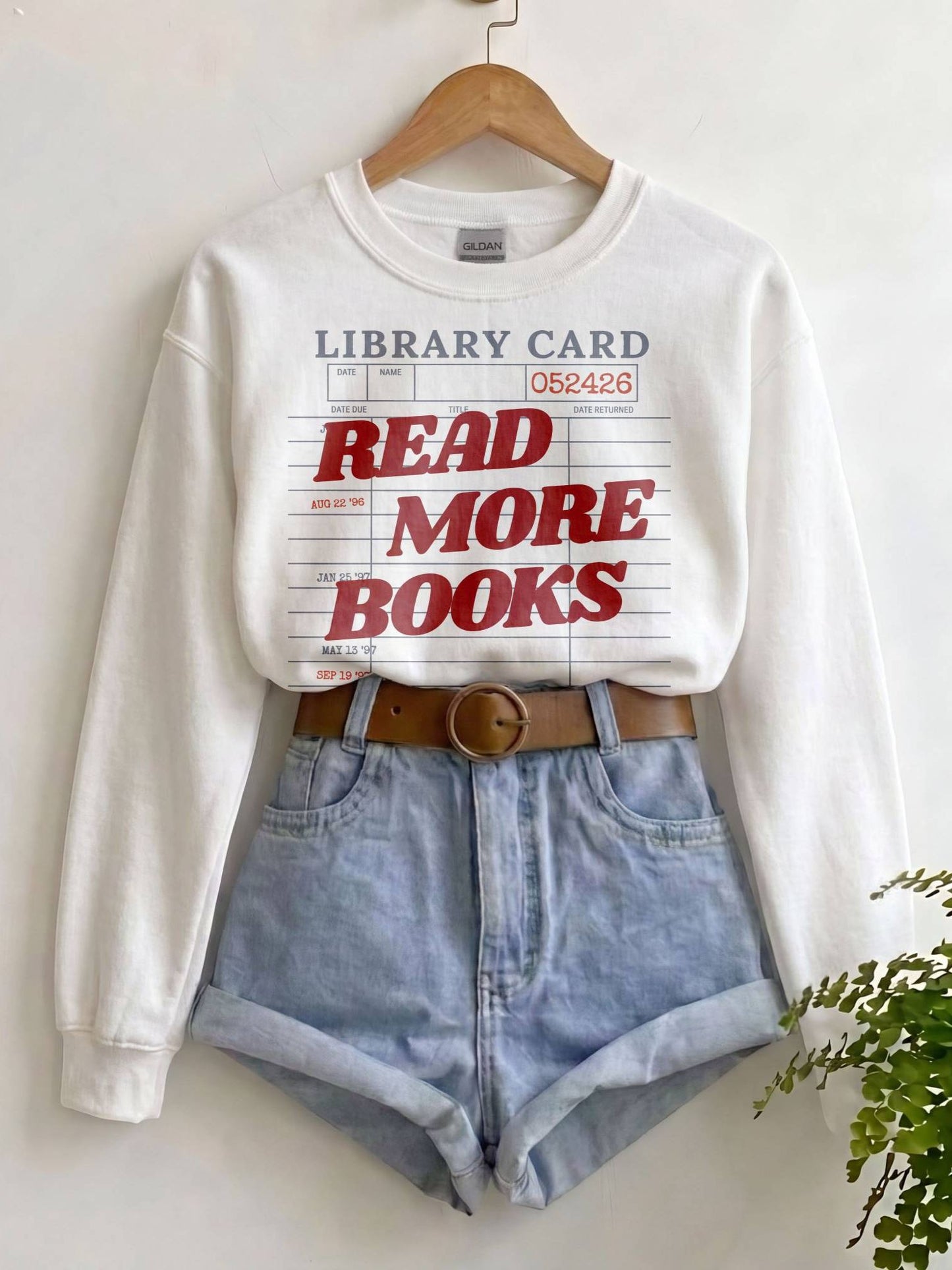 A white crewneck sweatshirt with a library card graphic that reads 'READ MORE BOOKS' in red, styled on a wooden hanger with light blue denim shorts and a brown belt against a white wall.