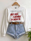 A white crewneck sweatshirt with a library card graphic that reads 'READ MORE BOOKS' in red, styled on a wooden hanger with light blue denim shorts and a brown belt against a white wall.