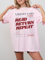 woman wearing a blossom pink Comfort Colors shirt featuring a vintage library card design with read return repeat text in red, oversized fit styled with biker shorts for a casual reader and librarian inspired look