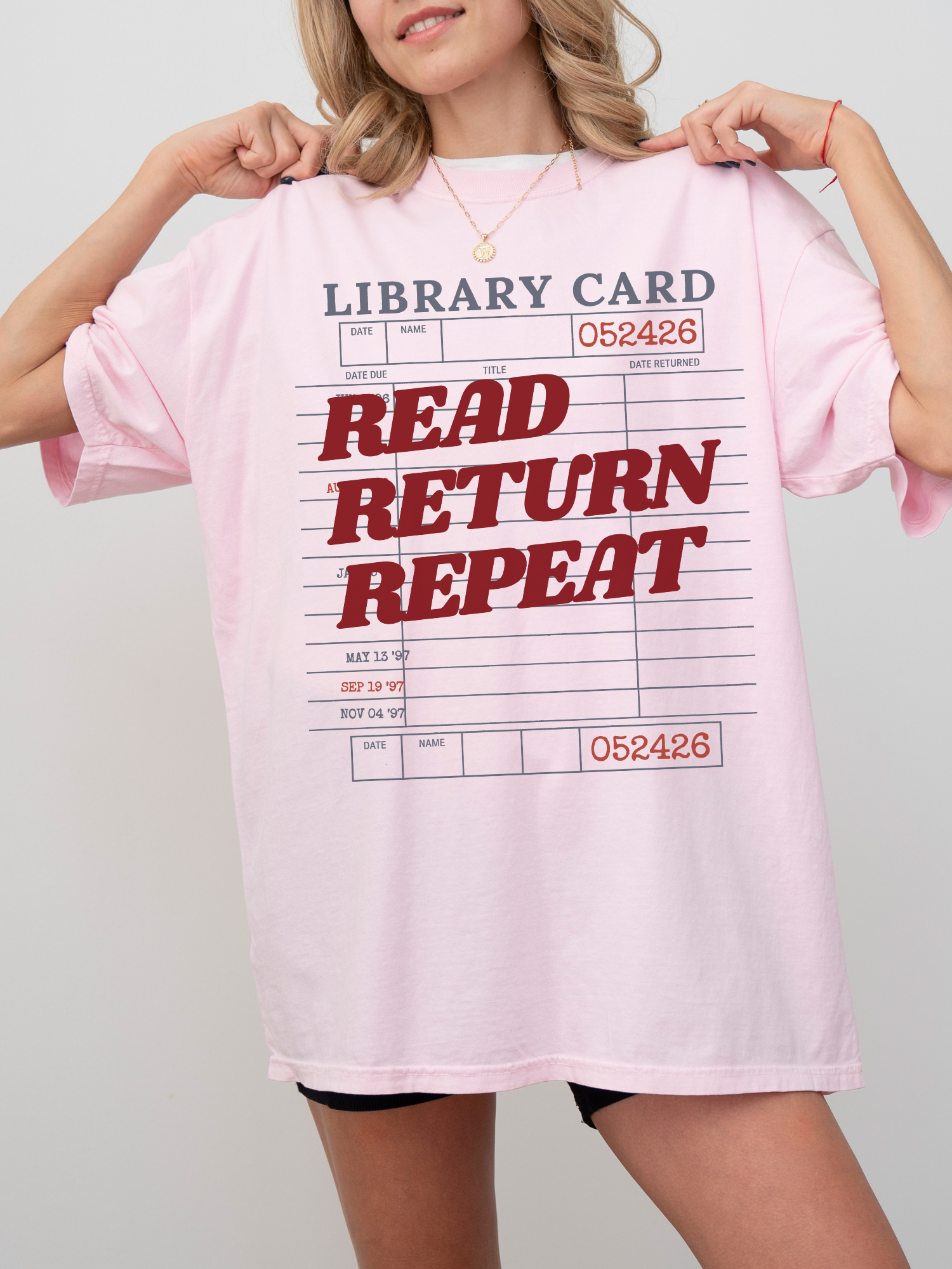 woman wearing a blossom pink Comfort Colors shirt featuring a vintage library card design with read return repeat text in red, oversized fit styled with biker shorts for a casual reader and librarian inspired look
