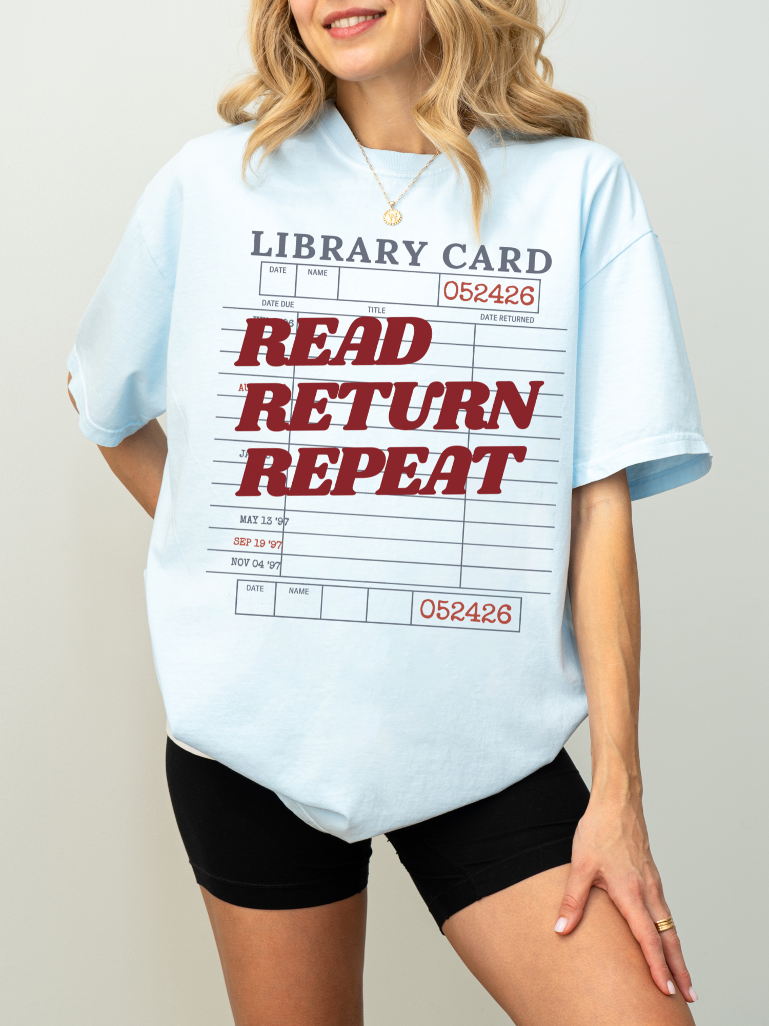 woman wearing a chambray blue Comfort Colors shirt featuring a library card inspired read return repeat graphic with red lettering, paired with black biker shorts for a relaxed bookworm outfit