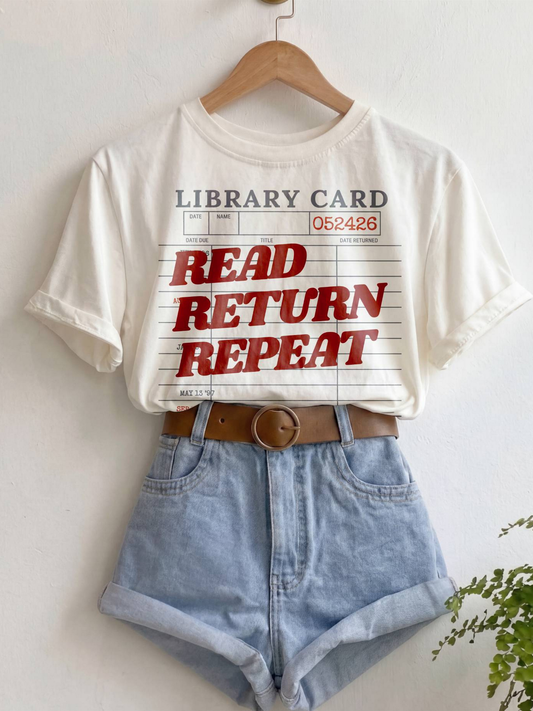 ivory Comfort Colors shirt hanging with rolled sleeves and a vintage library card graphic featuring the phrase read return repeat in red lettering, styled with denim shorts and a brown belt for a cozy bookish aesthetic