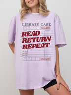 woman wearing an orchid Comfort Colors shirt featuring a retro library card graphic with read return repeat text in bold red letters, oversized book lover tee styled with biker shorts and gold jewelry