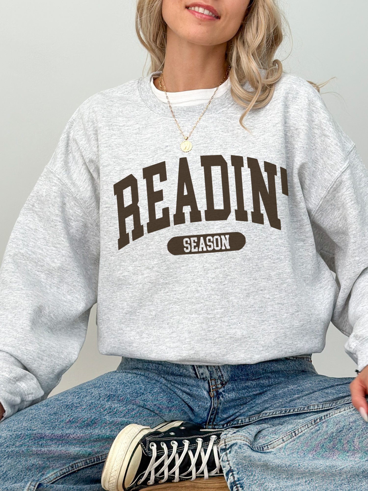 Ash grey crewneck with Readin' Season in bold brown varsity font, paired with classic blue jeans and black Converse