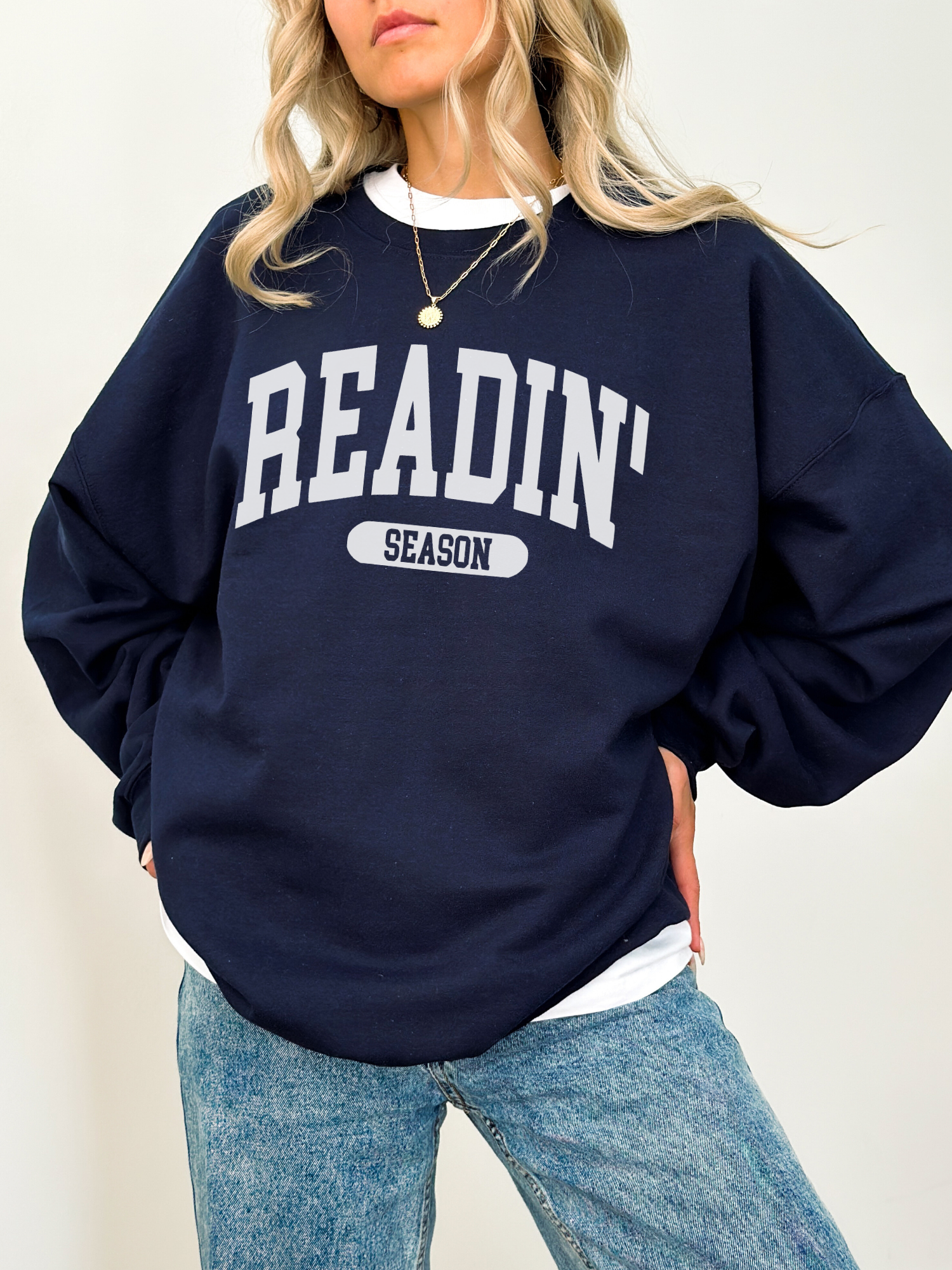 Navy sweatshirt with Readin' Season in light grey varsity text, worn over a white shirt and styled with relaxed denim
