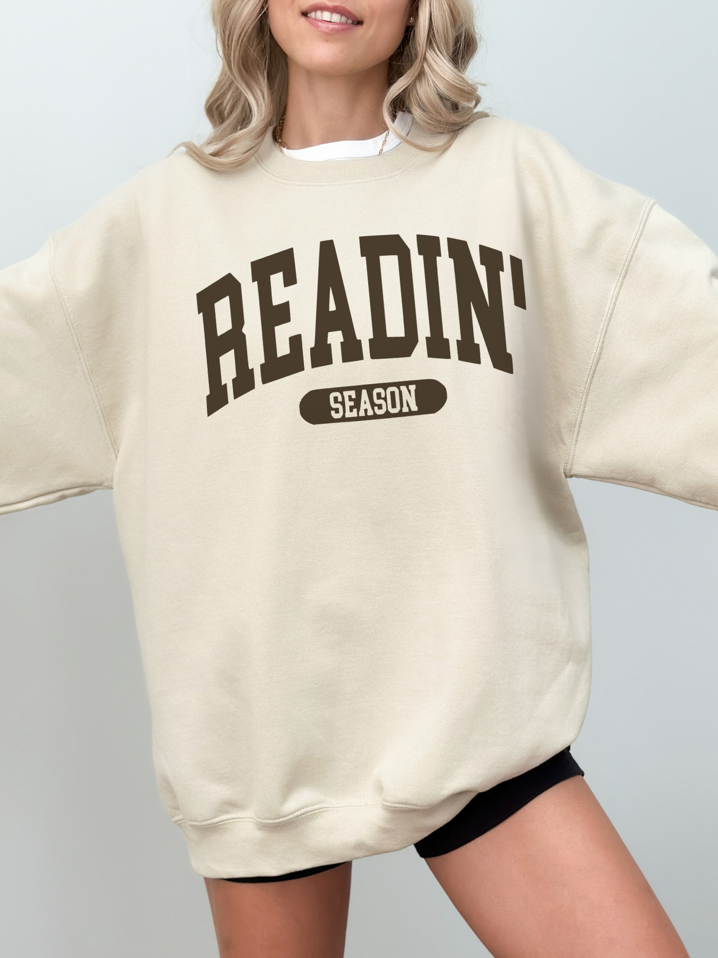 Cream colored sweatshirt featuring Readin' Season text in chocolate brown, styled oversized on a smiling woman with wavy blonde hair
