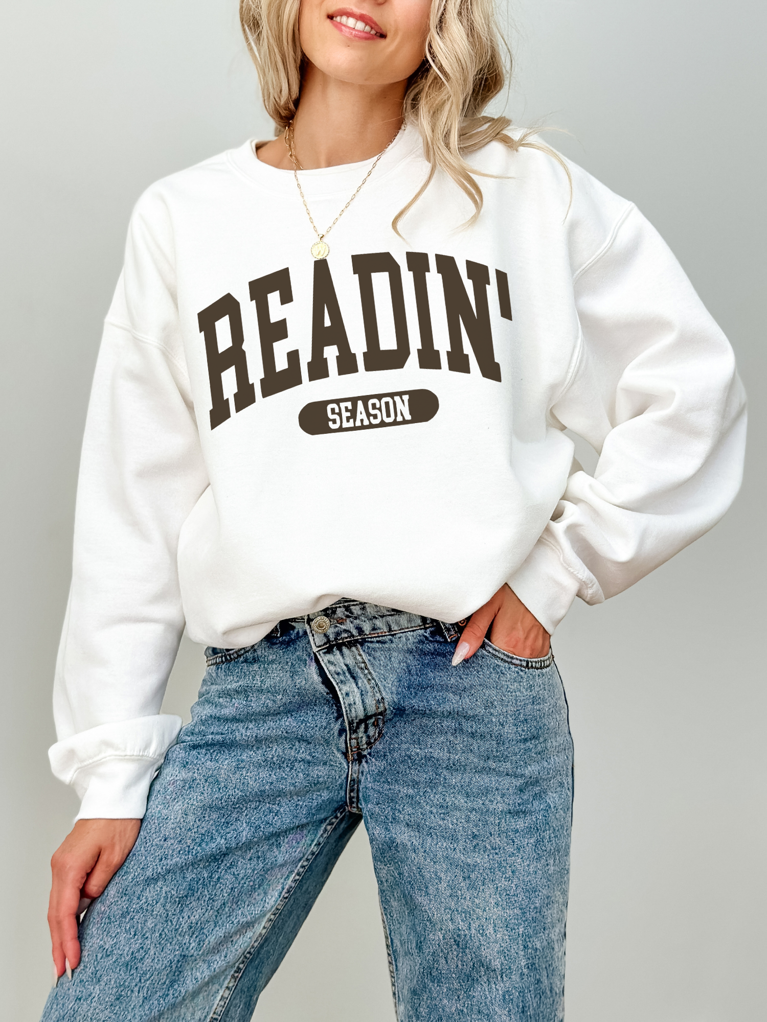 White sweatshirt with bold varsity-style Readin' Season text in dark brown, worn by a blonde woman with light-wash jeans and a gold coin necklace