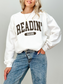 White sweatshirt with bold varsity-style Readin' Season text in dark brown, worn by a blonde woman with light-wash jeans and a gold coin necklace