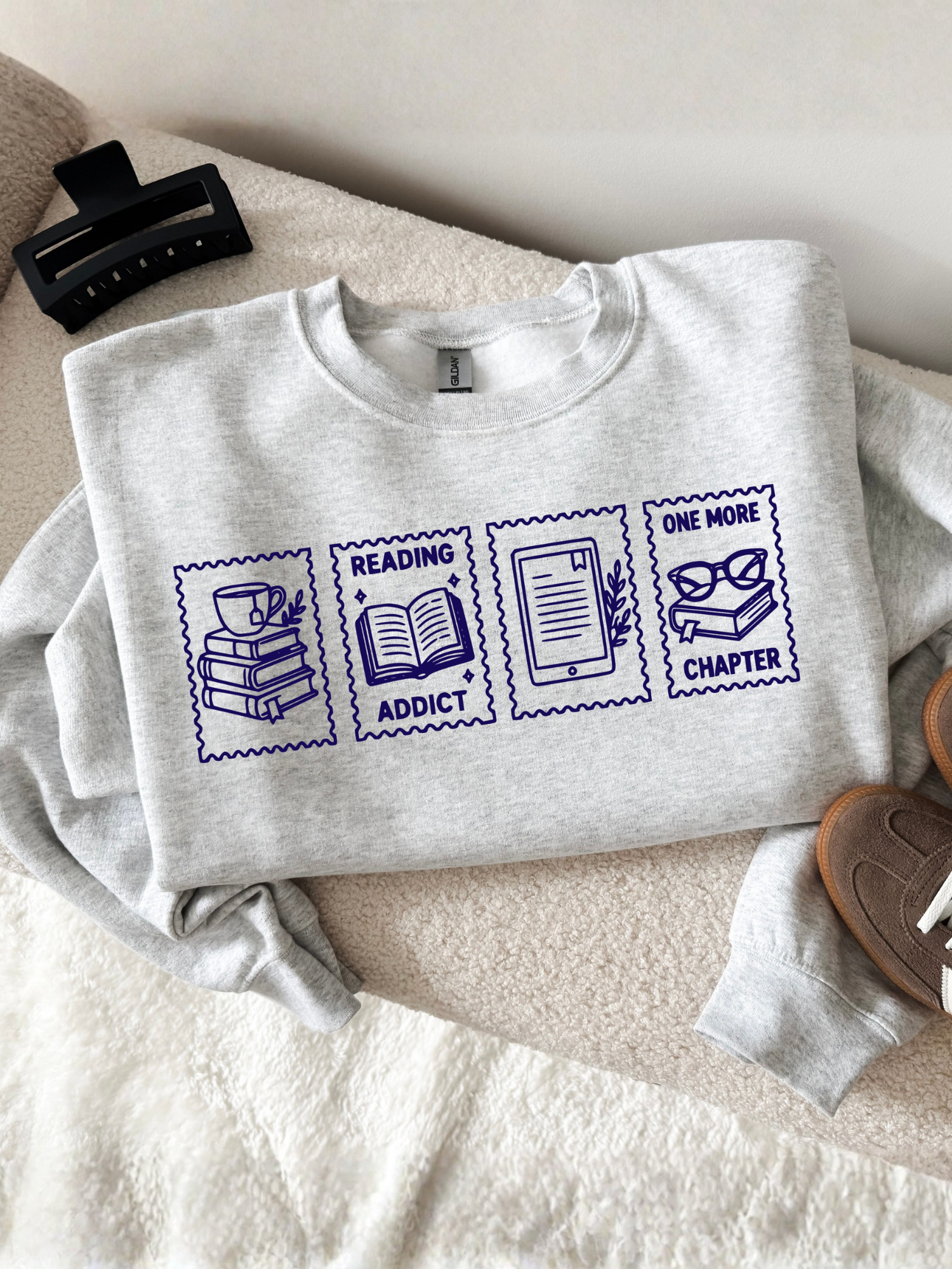 folded ash gray sweatshirt displayed on a cozy chair featuring reading addict one more chapter stamp graphic with books coffee open book e reader and glasses illustrations