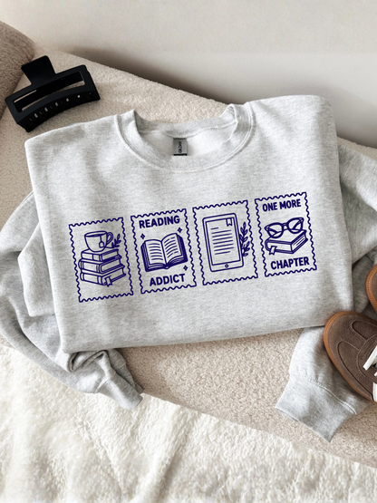 folded ash gray sweatshirt displayed on a cozy chair featuring reading addict one more chapter stamp graphic with books coffee open book e reader and glasses illustrations