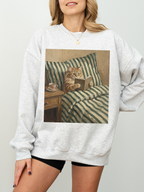 Light grey sweatshirt featuring a painted style illustration of a cat reading a book in bed next to a cup of tea, worn by a model indoors. The soft neutral styling creates a relaxed and cozy aesthetic, perfect for book lovers, readers, and cat lovers who enjoy comfortable and literary inspired clothing.