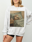 Light grey sweatshirt featuring a painted style illustration of a cat reading a book in bed next to a cup of tea, worn by a model indoors. The soft neutral styling creates a relaxed and cozy aesthetic, perfect for book lovers, readers, and cat lovers who enjoy comfortable and literary inspired clothing.