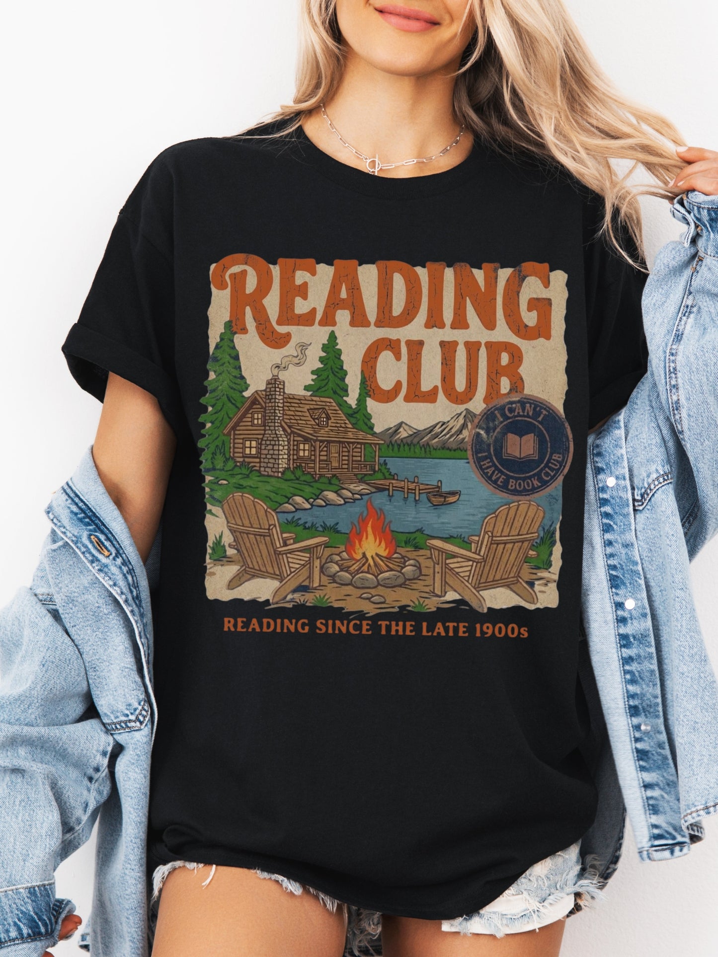A woman wearing a black graphic t-shirt with a vintage-style 'Reading Club' print, styled with an open light-wash denim jacket.