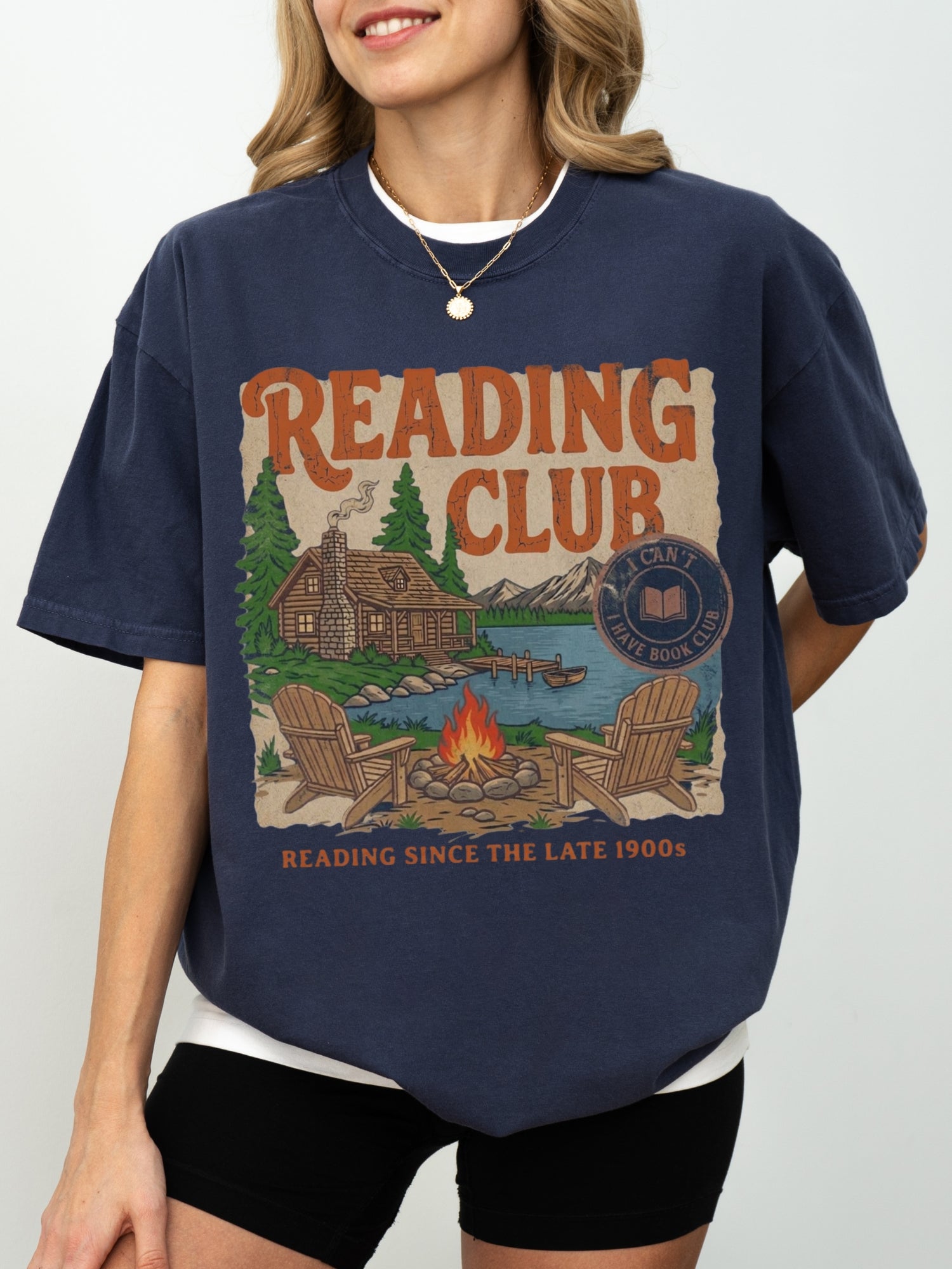 A woman wearing a navy blue graphic t-shirt with a vintage-style 'Reading Club' print on the front, paired with black shorts against a light gray studio background.