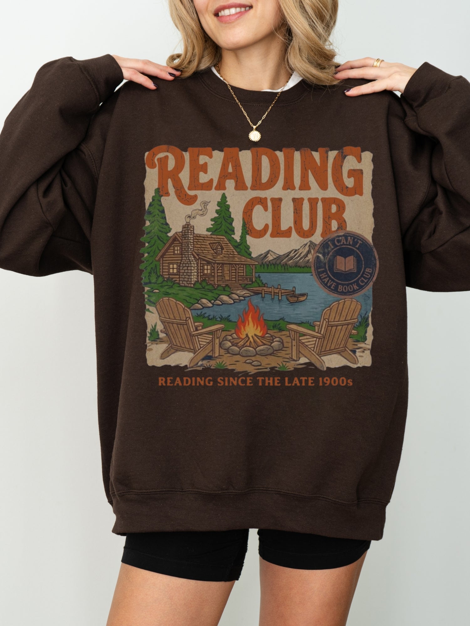A woman wearing a chocolate brown oversized sweatshirt with a vintage-style 'Reading Club' graphic on the front, paired with black shorts against a light gray studio background.