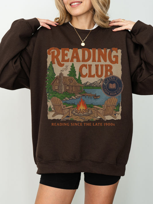 A woman wearing a chocolate brown oversized sweatshirt with a vintage-style 'Reading Club' graphic on the front, paired with black shorts against a light gray studio background.