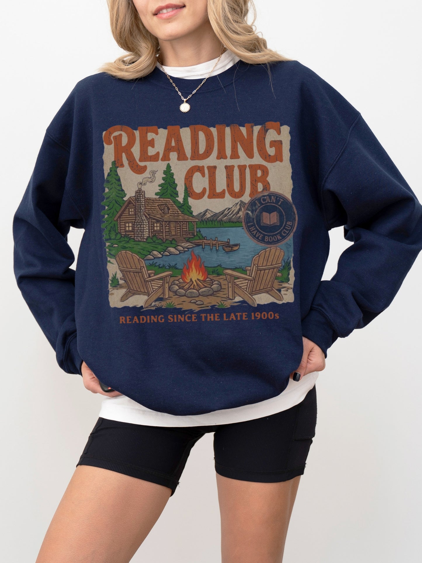A woman wearing a navy blue crewneck sweatshirt with a large vintage-style graphic on the front that reads 'READING CLUB' above an illustration of a cabin, lake, campfire, and chairs, paired with black shorts against a light gray studio background.