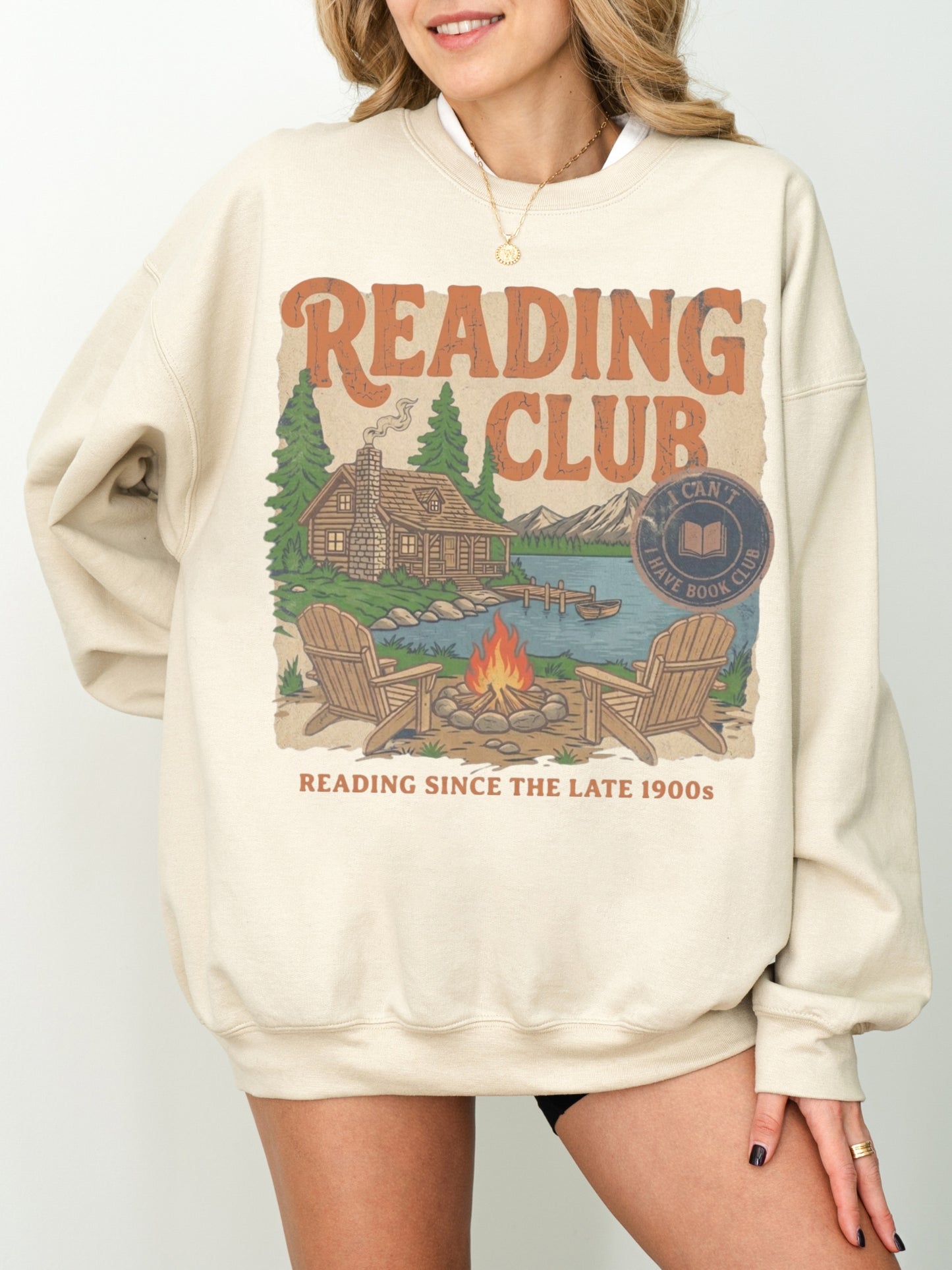 A woman wearing an oversized sand-colored sweatshirt with a vintage-style 'Reading Club' graphic on the front, featuring a cabin, lake, trees, and campfire illustration.