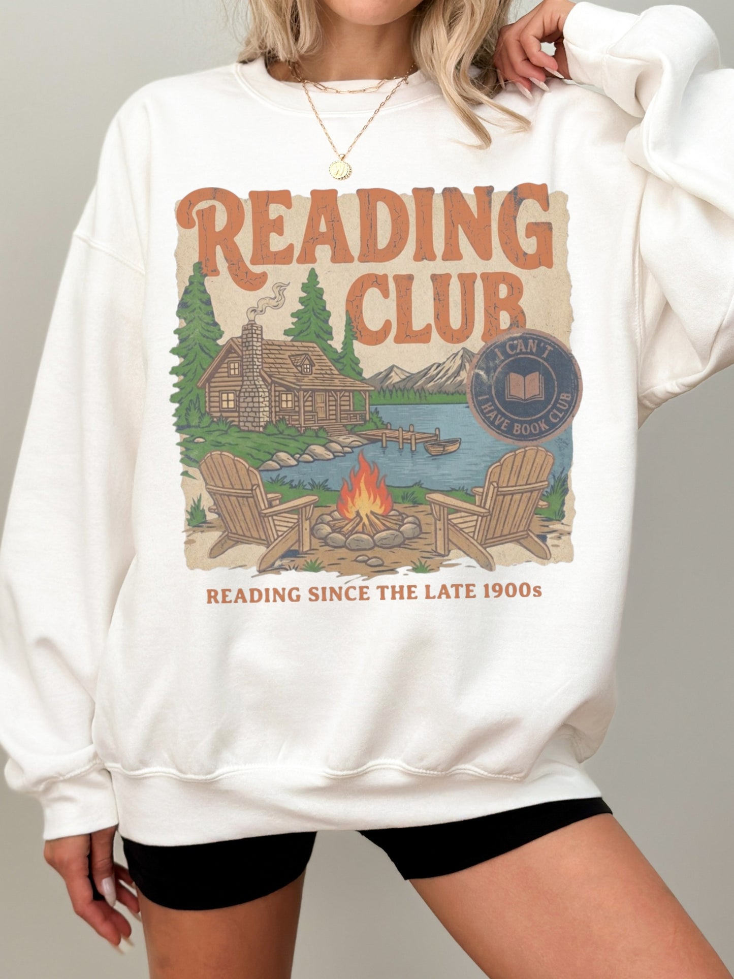 A woman wearing an oversized white crewneck sweatshirt with a vintage-style 'Reading Club' graphic on the front, paired with black shorts, against a solid gray studio background.