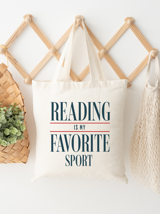Canvas tote bag with reading is my favorite sport printed in bold serif text, hanging on a wooden wall rack next to woven baskets and greenery. The neutral aesthetic styling creates a cozy bookish home vibe, making it perfect for readers, bookstore trips, and everyday errands while showing off a love for reading.