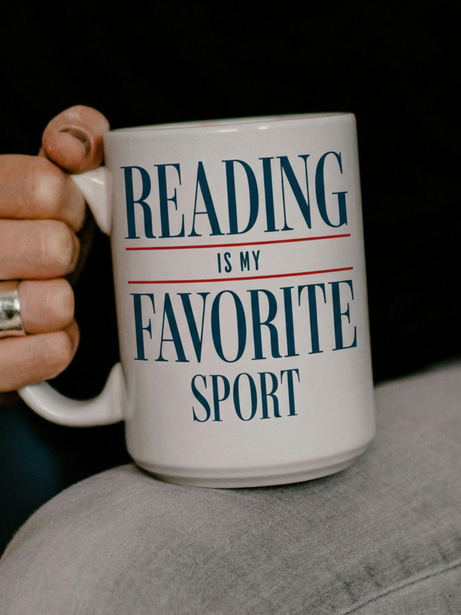 White ceramic mug with reading is my favorite sport printed in bold text, held in one hand in a darker cozy setting. The lifestyle photo highlights a relaxed reading vibe, perfect for book lovers enjoying coffee or tea, with soft shadows and casual styling that emphasizes a calm and comfortable reading atmosphere.