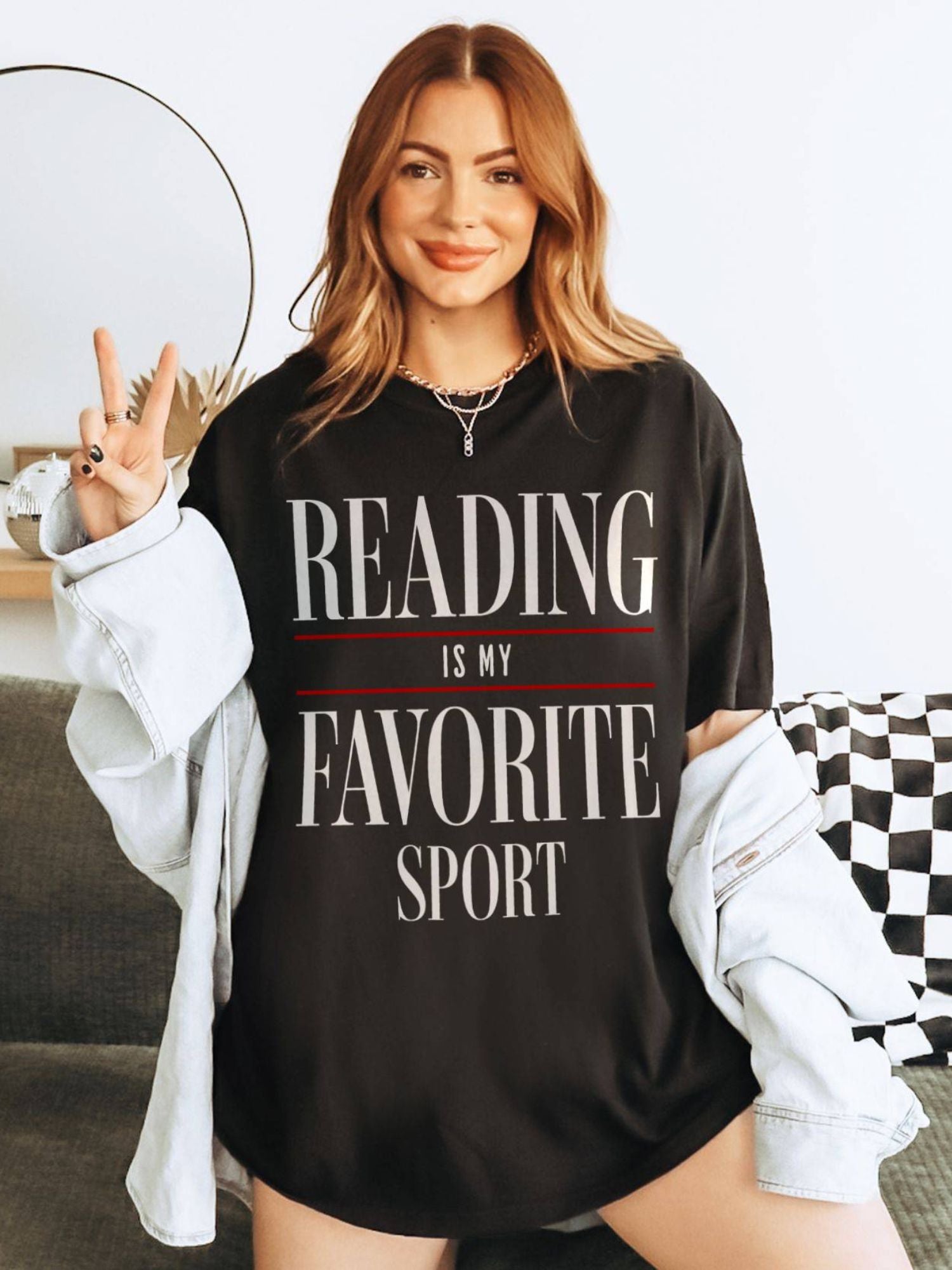 Woman wearing oversized "Reading Is My Favorite Sport" graphic tee in black with bold text, smiling casually and giving a peace sign with her hand

