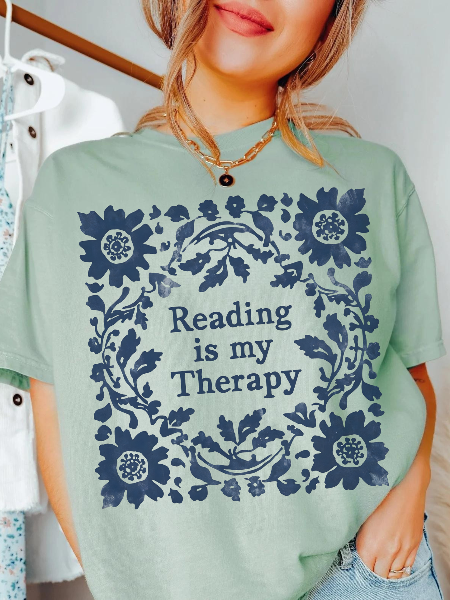 A woman wearing a bay green t-shirt with a navy blue floral wreath graphic and the text 'Reading is my Therapy' printed on the front.