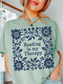 A woman wearing a bay green t-shirt with a navy blue floral wreath graphic and the text 'Reading is my Therapy' printed on the front.