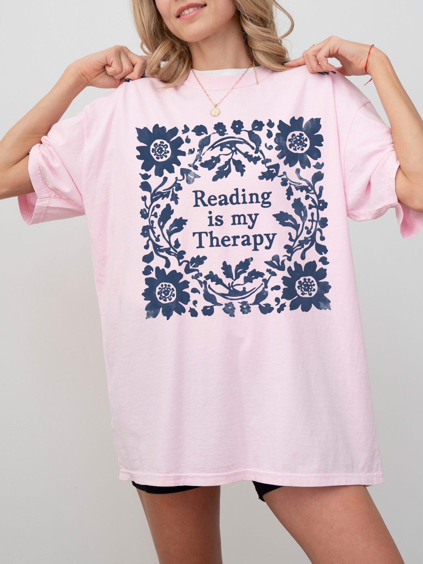 A woman wearing an oversized blossom t-shirt with a navy blue floral border and the text 'Reading is my Therapy' printed on the front, set against a plain light gray background.