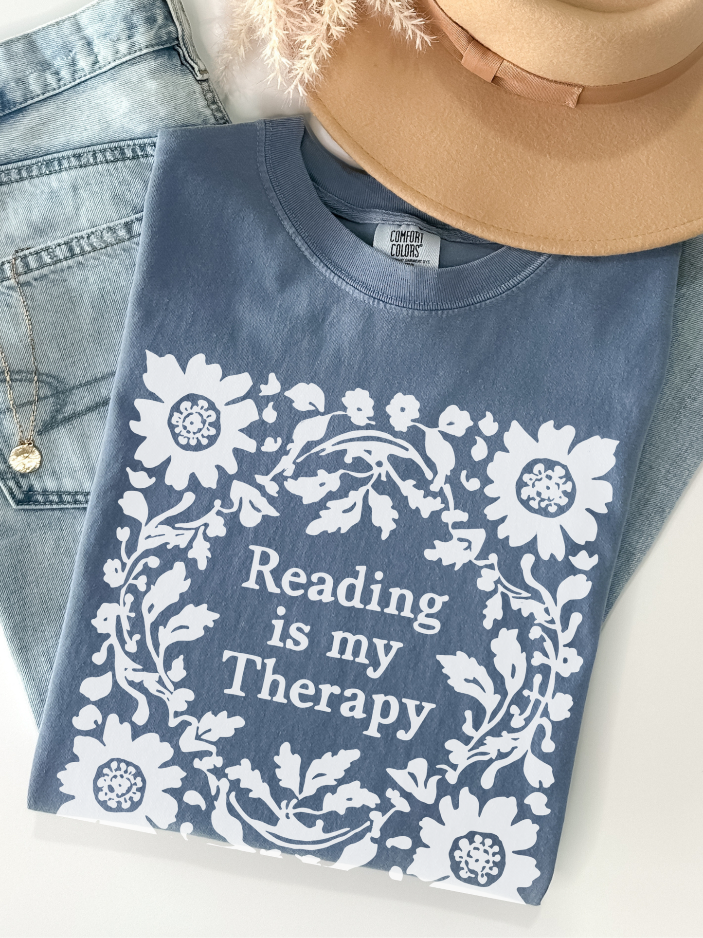 A folded blue jean t-shirt with a white floral wreath graphic and the text 'Reading is my Therapy' in white, styled with a tan hat, blue jeans, and a gold necklace on a white background.
