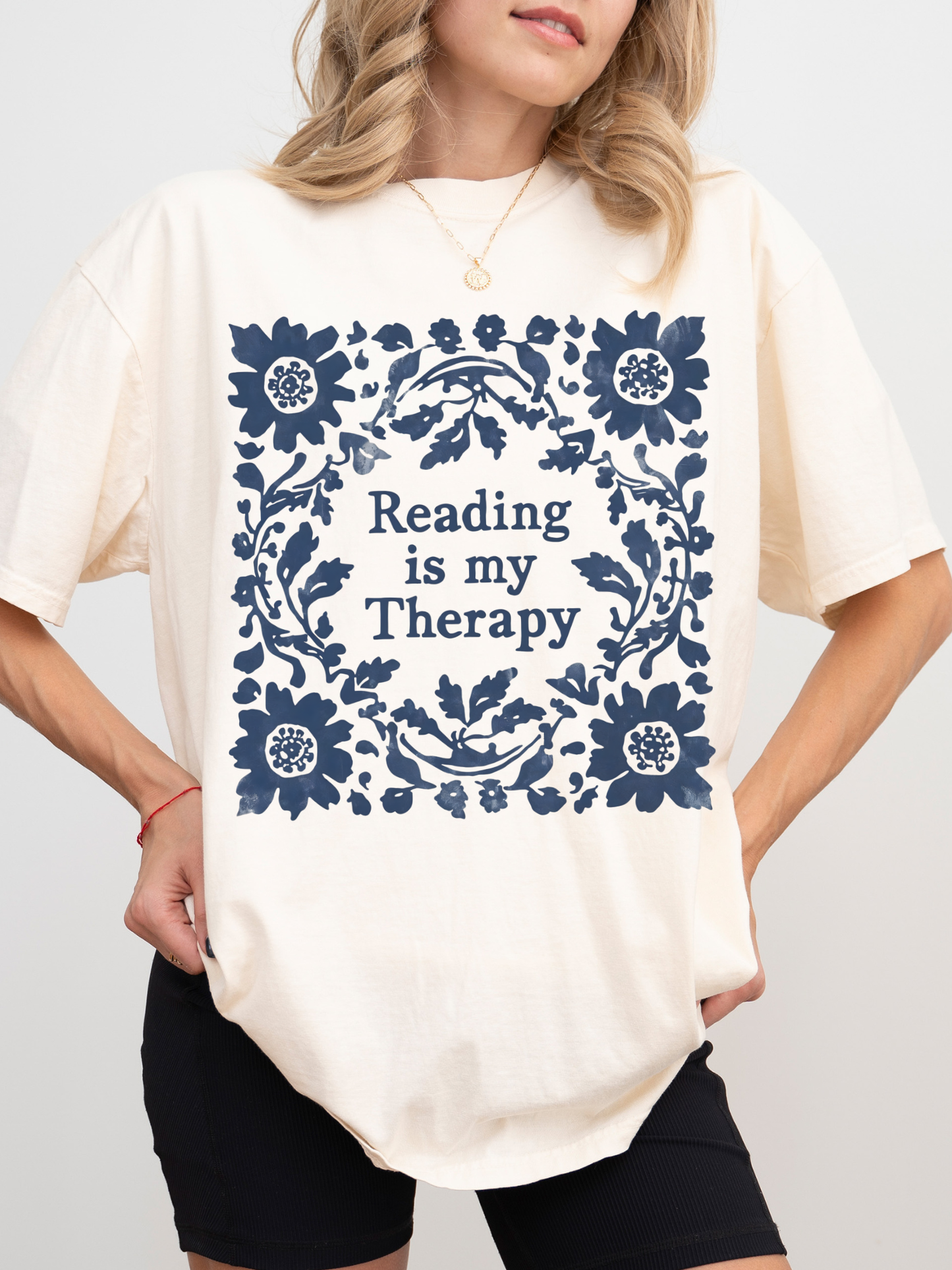 A woman wearing an oversized ivory-colored t-shirt with a navy blue floral border and the text 'Reading is my Therapy' printed on the front, paired with black shorts.