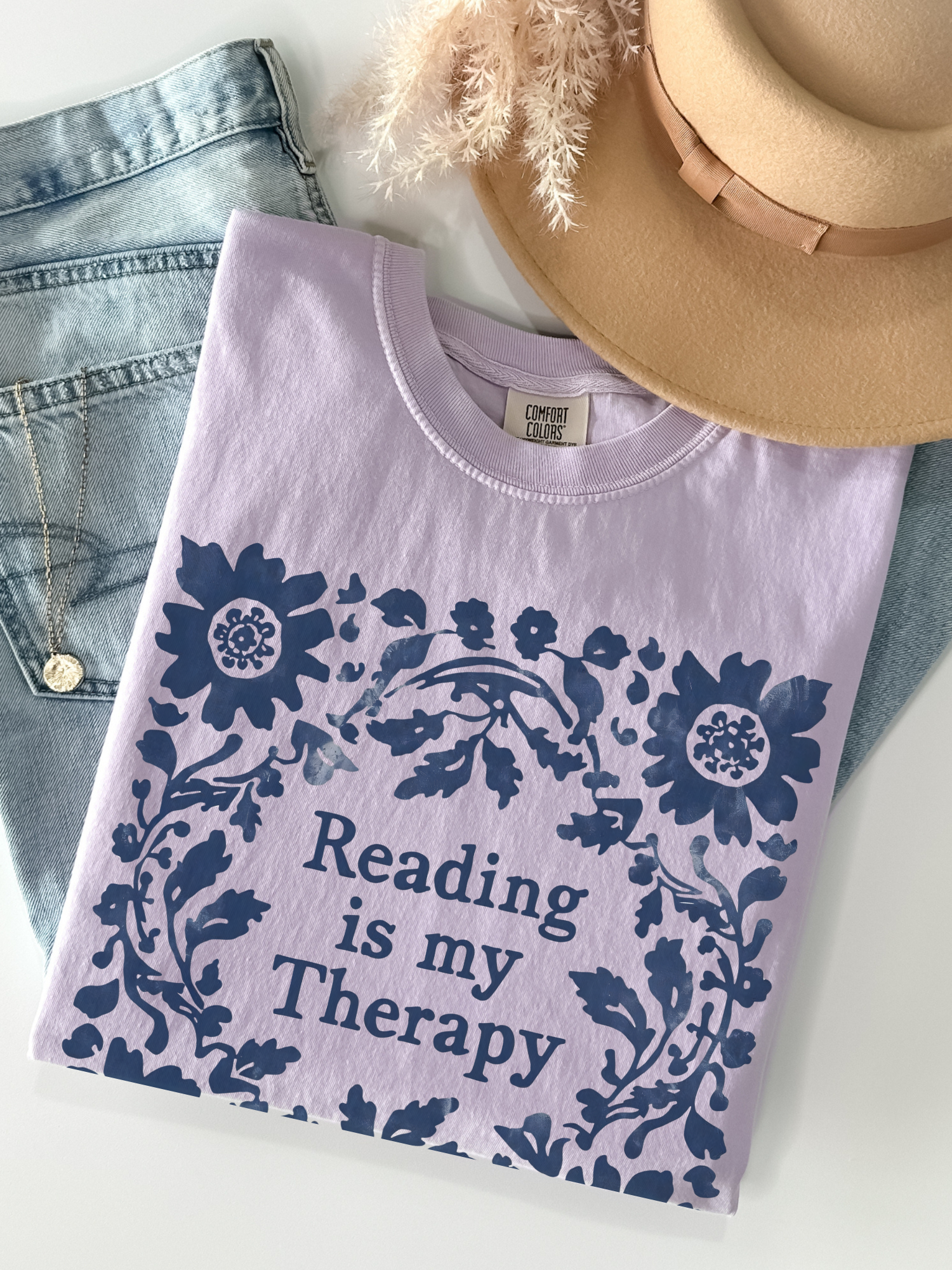 A folded orchid crewneck t-shirt with a navy blue floral border and the text 'Reading is my Therapy' in the center, styled in a flat lay with blue jeans, a tan hat, and a gold necklace on a white background.