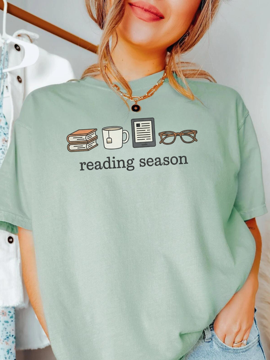 Woman wearing a Bay Comfort Colors shirt with Reading Season design featuring books, tea mug, Kindle, and glasses, styled with denim jeans and gold necklace