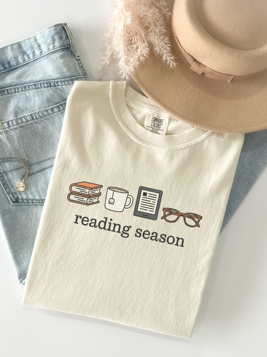 Flat lay of ivory Reading Season shirt with books, mug, Kindle, and glasses, styled with light wash jeans and a felt hat