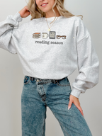 Ash grey reading season crewneck with book, tea, Kindle, and glasses icons, styled with high-waisted jeans and layered gold necklace — soft and cozy sweatshirt for bookworms.