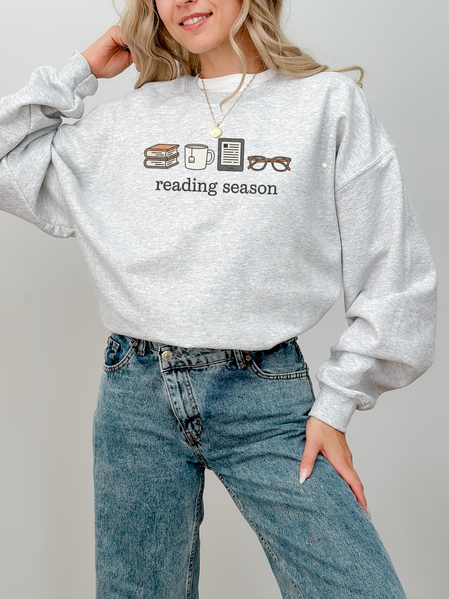 Ash grey reading season crewneck with book, tea, Kindle, and glasses icons, styled with high-waisted jeans and layered gold necklace — soft and cozy sweatshirt for bookworms.