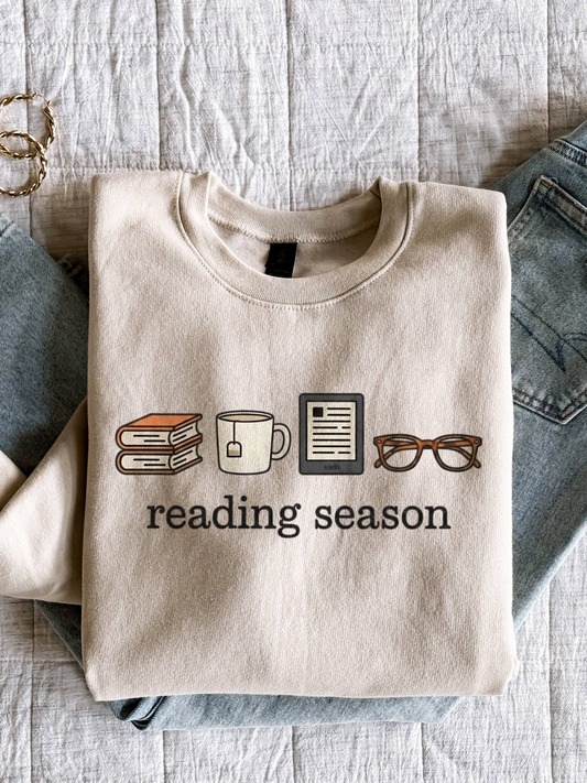 Sand-colored reading season sweatshirt folded neatly on a bed with jeans and gold hoop earrings, featuring cozy bookish icons — perfect flat lay for fall reading vibes and warm neutral tones.