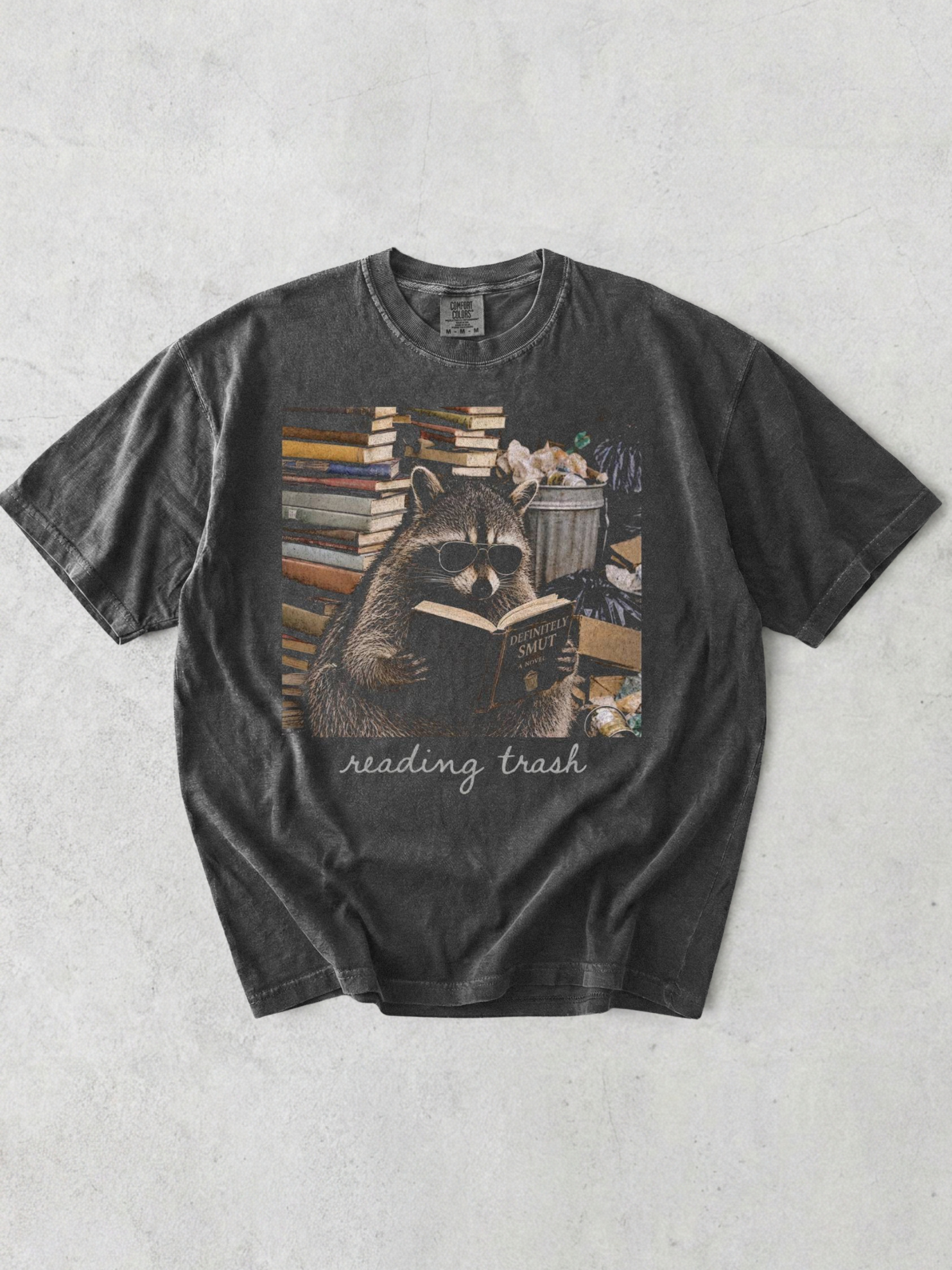 pepper comfort colors reading trash shirt featuring raccoon wearing sunglasses reading smut novel surrounded by books and trash cans