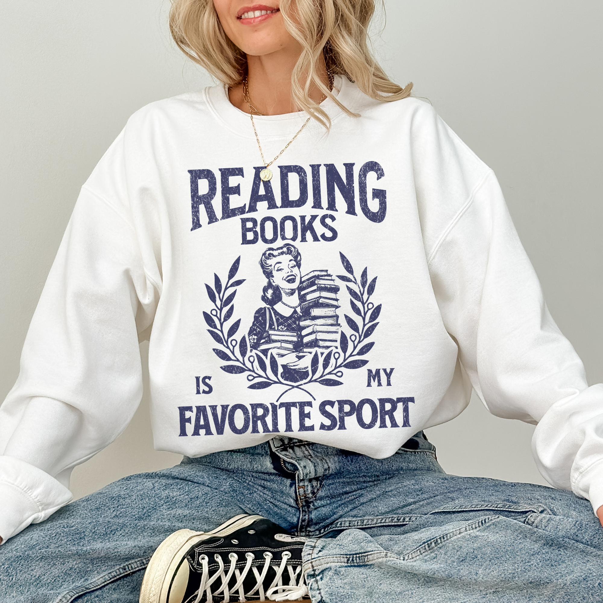 woman sitting cross-legged in a white sweatshirt that says reading books is my favorite sport with navy graphic