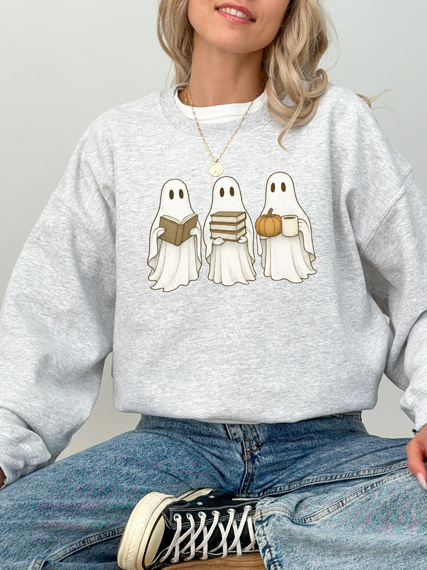 woman wearing an ash grey sweatshirt with three cute cartoon ghosts holding a book, a stack of books, and a pumpkin mug
