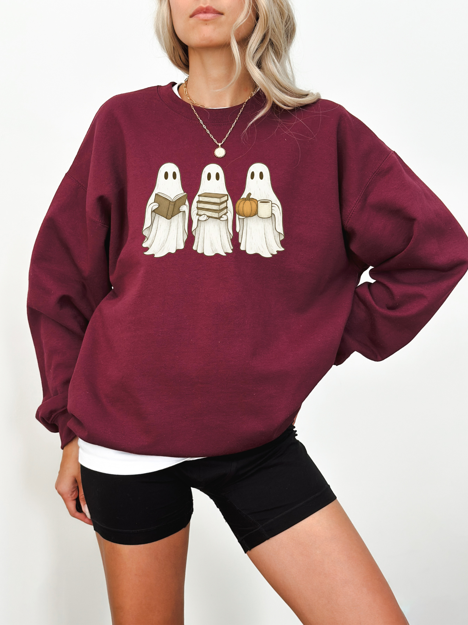 woman wearing a maroon sweatshirt with three cute ghosts holding a book, stacked books, and a pumpkin mug