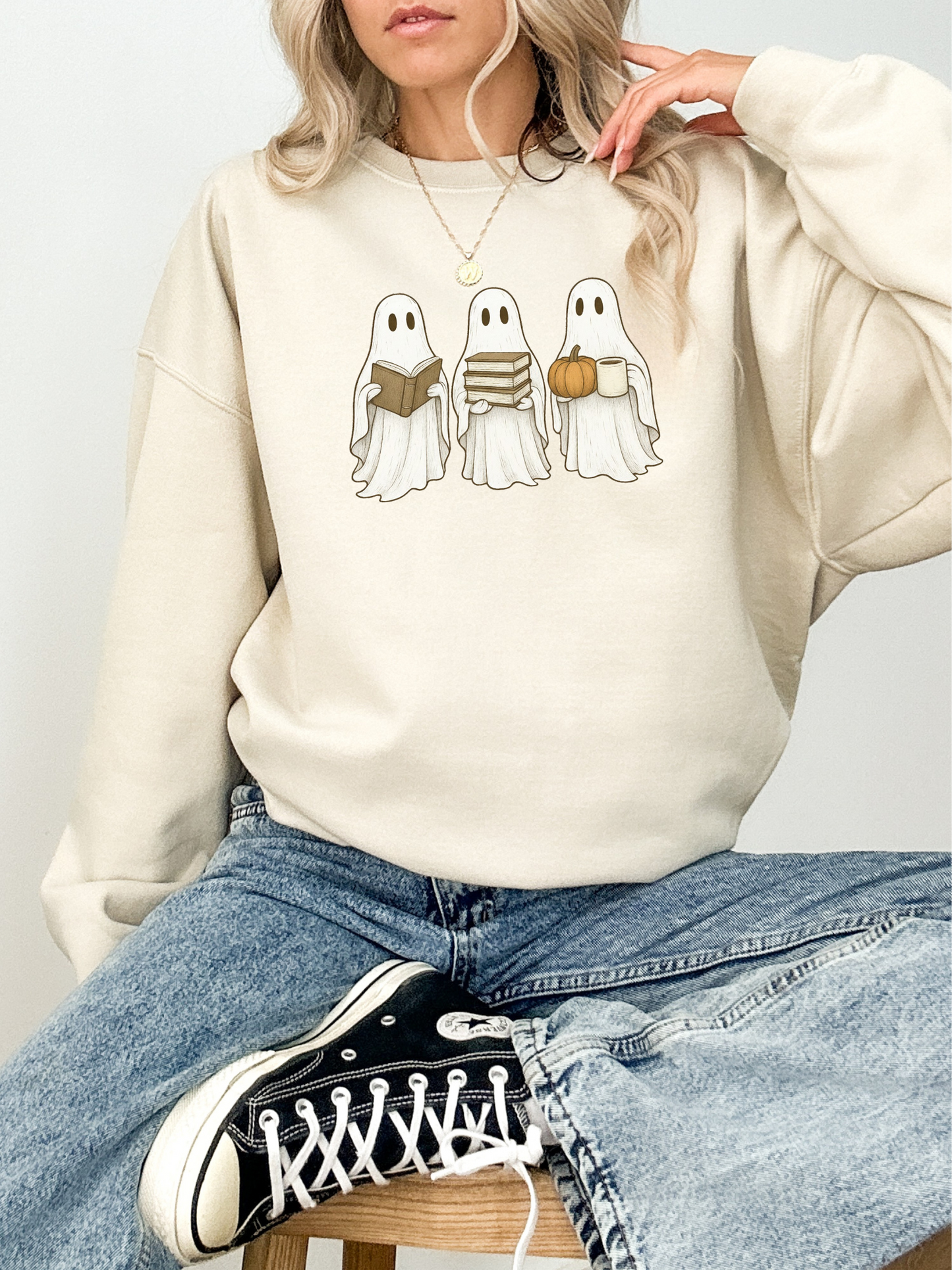 woman in a sand-colored sweatshirt with cartoon ghosts holding fall-themed bookish items, styled for Halloween
